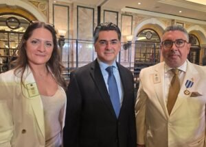 WhatsApp Image 2026-07-26 at 15356 PM1 - عروبة الاخباري | Oroba News Three professionals posing for a photo at an indoor formal event; two wear light beige blazers with badges, one in a dark suit and blue tie in the middle.