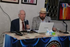 Elderly man in a suit speaks into a microphone at a table, while a bearded man wearing a hat sits with a white Toshiba laptop beside him.