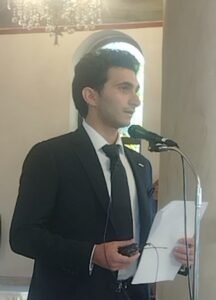 WhatsApp Image 2026-07-28 at 115432 AM - عروبة الاخباري | Oroba News Young man in a dark suit and tie speaks into a microphone while holding papers at a formal event.