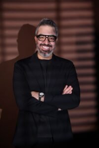 Smiling man with salt‑and‑pepper hair and beard, wearing a black blazer and glasses, arms crossed in front of a striped background.