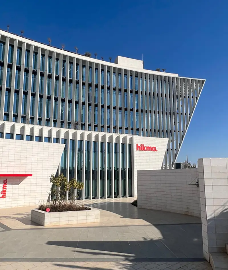 amman-hq-mena-jordan - عروبة الاخباري | Oroba News Facade of a modern Hikma building with tall glass windows and vertical fins, white tiled walls, blue sky, and a small landscaped planter in the foreground.