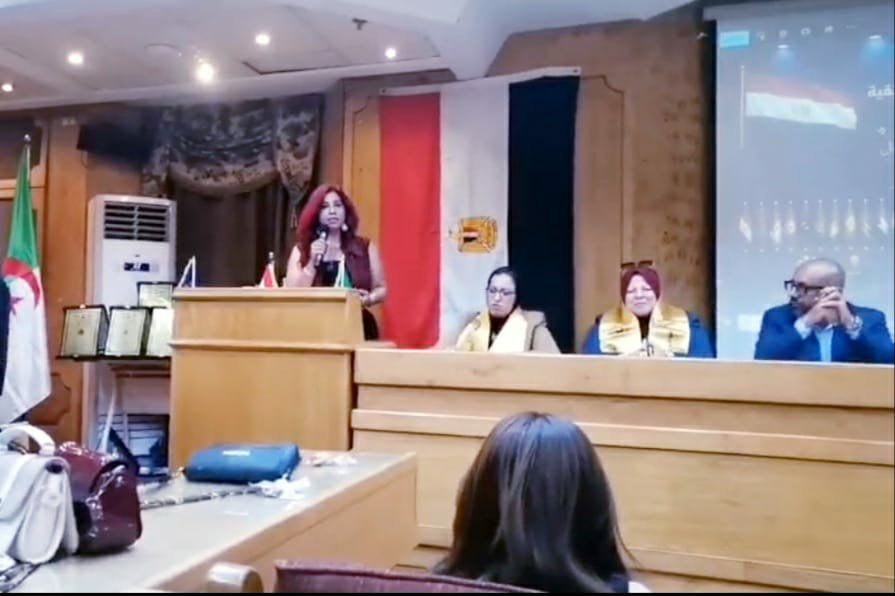Woman with red hair speaks at a wooden podium during a panel discussion, flanked by two seated panelists, with flags and a projector screen in the background.