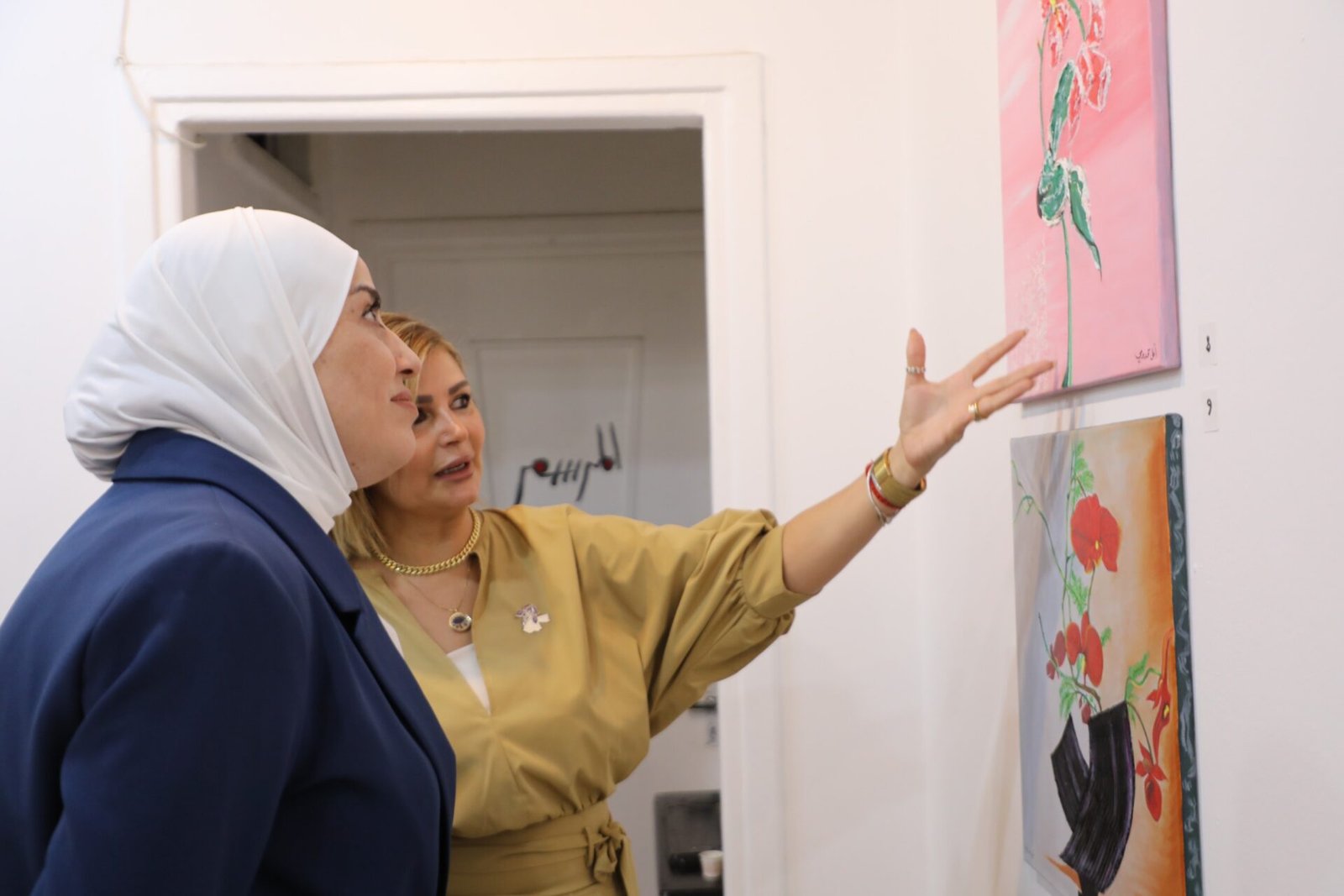 h6ZwySXOf5noV3MQ5Jj6DQA4PrZnSEHDzCuX3zA2 Two women in a gallery observe colorful paintings on the wall, one in a white hijab and blue blazer, the other gesturing toward the art.