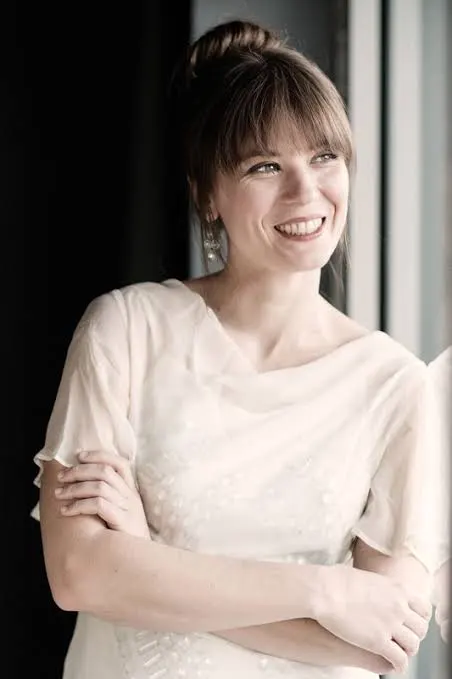 images565588000 Smiling woman in a light dress standing by a window, arms loosely crossed.
