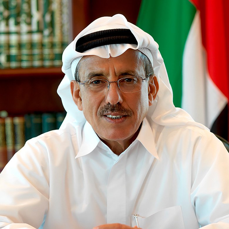Middle-aged man in a white thobe and ghutra, wearing glasses, seated at a desk with a flag behind him.