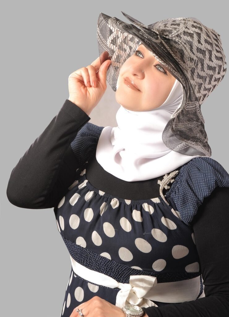 Woman wearing a white hijab and navy polka-dot dress, tipping a wide-brim patterned hat.