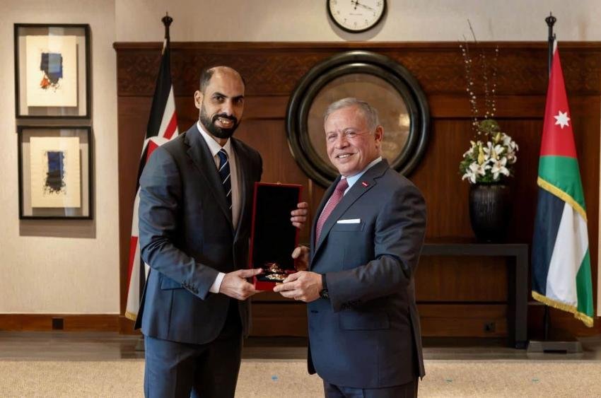 Two men in suits exchange a red award plaque in a formal office, with flags and a clock behind them.—award ceremony scene