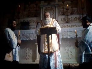1-3-300x225 - عروبة الاخباري | Oroba News Old priest in liturgical robes delivers a sermon at a pulpit, flanked by two assistants holding candles in a church interior.