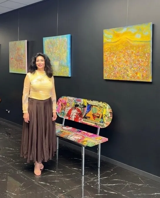 1_news_1787842975 - عروبة الاخباري | Oroba News Woman in a gallery stands beside a colorful graffiti-style bench as three abstract paintings hang on a dark wall behind her.