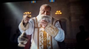 2-2-300x167 - عروبة الاخباري | Oroba News Bearded priest in white and gold liturgical vestments blessing with raised right hand during a church service