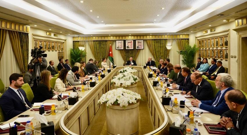 A formal international meeting around a long oval conference table, with officials, cameras, and a central flower arrangement in a government-style room.