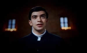 4-2-300x184 - عروبة الاخباري | Oroba News Young priest in a black clerical shirt with a white collar, lit softly in a church setting.