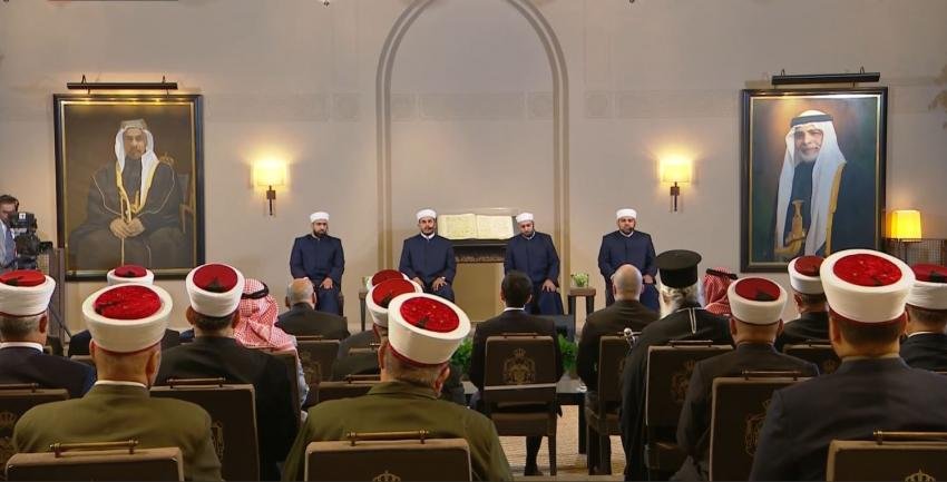 Five men in dark suits sit on a stage before an audience wearing red-and-white head turbans in a ceremonial hall with two large portraits on the walls.