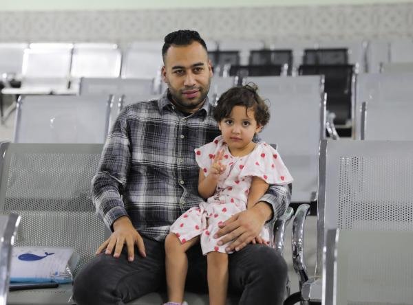 Father and young daughter sit together in a row of gray waiting chairs, looking at the camera.
