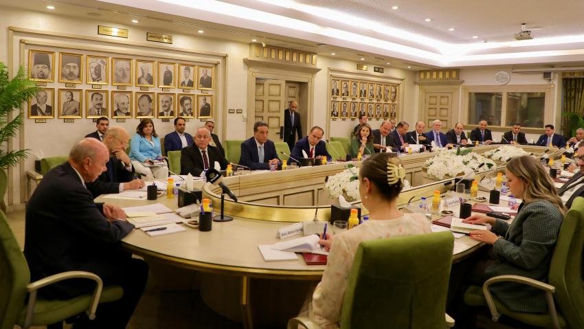 Formal boardroom meeting with dozens of attendees seated around a large oval table, microphones, papers, and flowers, portraits lining the walls.