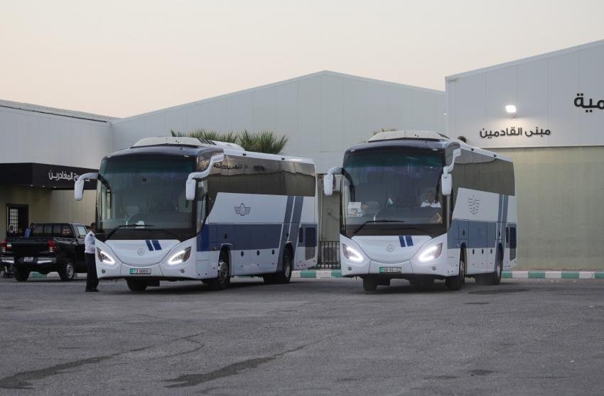 Two white and blue passenger buses parked side by side in a parking lot near a large warehouse-like building with Arabic signage in the background.