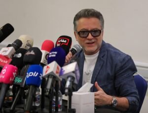 550023_0_1785519521 - عروبة الاخباري | Oroba News Man with glasses in a blue blazer speaks into a handheld microphone at a press conference, surrounded by many microphones from news outlets on a table.
