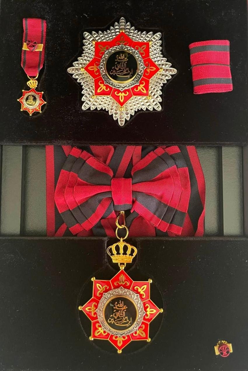 Decorative medal set: red and black striped ribbons, a star-shaped red-and-silver medal, a gold crown charm, and a matching medallion with Arabic inscription on a black display case.