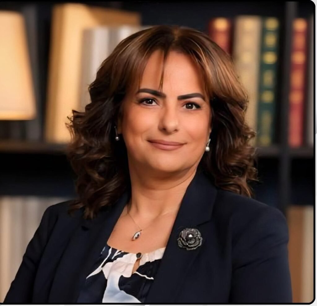 5f0ded2e-c9b9-4db8-b695-f5284e66f8f0 - عروبة الاخباري | Oroba News Portrait of a professional woman with brown hair in a navy blazer, library backdrop behind her, smiling subtly with pearl earrings.