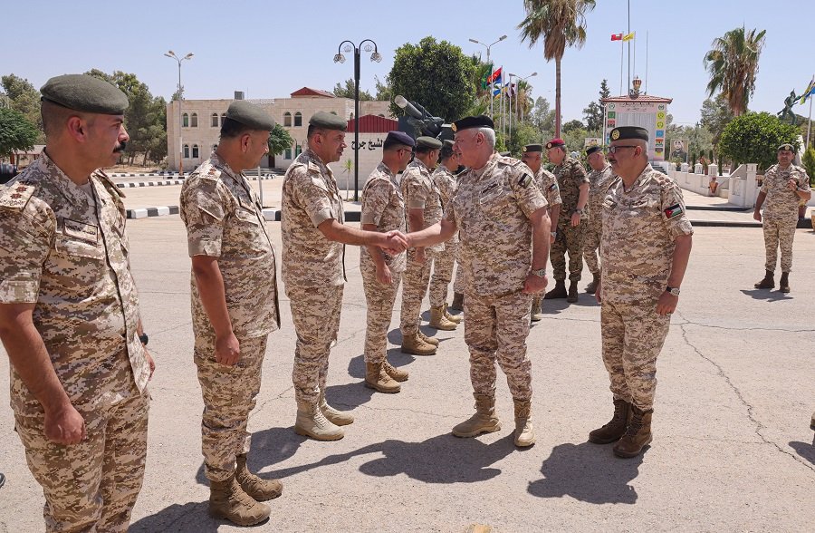 639214429532938691 Military officers in desert camouflage shake hands in a line during a ceremony on a sunlit base, with flags, palm trees, and a monument in the background.