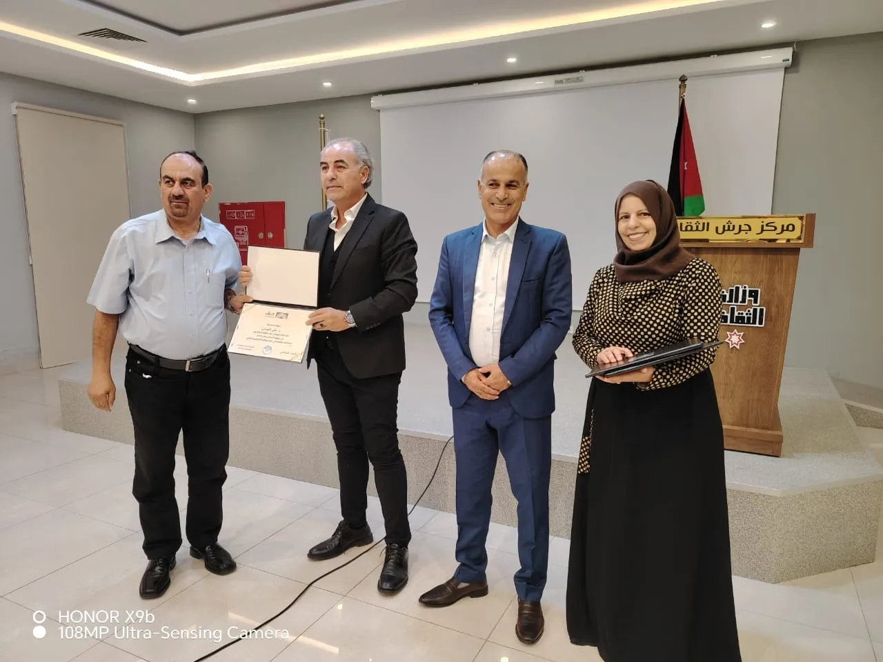 6a73a84475a56 - عروبة الاخباري | Oroba News Four people on a stage: a man in a light blue shirt hands a certificate to a man in a black suit while others watch, a podium with Arabic text and a Jordanian flag behind them.