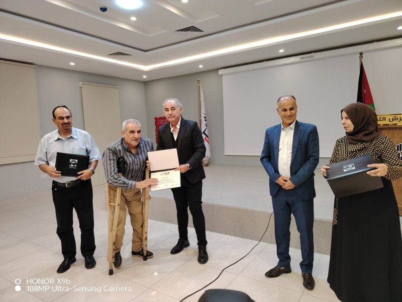 6a73a844b16bb - عروبة الاخباري | Oroba News Five people on a stage during an awards ceremony; a man with crutches receives a certificate from a suited presenter while others hold folders or boxes nearby.