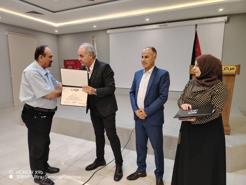 6a73a844cd640 - عروبة الاخباري | Oroba News Award ceremony: a man in a black suit presents a certificate to another man while two colleagues watch in a conference room with a flag in the background.