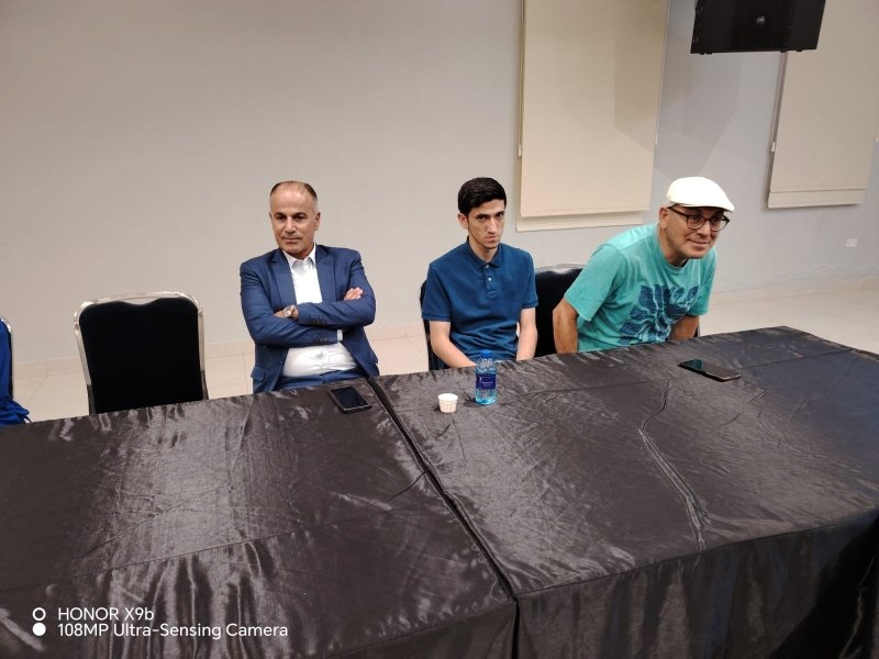 6a73a84503852 - عروبة الاخباري | Oroba News Three men sit at a long black table in a meeting room; left in a blue suit with arms crossed, middle in a blue polo, right in a teal shirt and white cap leaning forward with a phone and water bottle on the table beneath speakers.