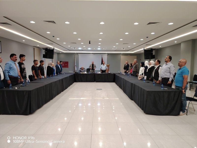6a73a8452fd58 - عروبة الاخباري | Oroba News People stand along a U-shaped conference table in a spacious meeting room, facing a central panel at the far end; water bottles line the tables.