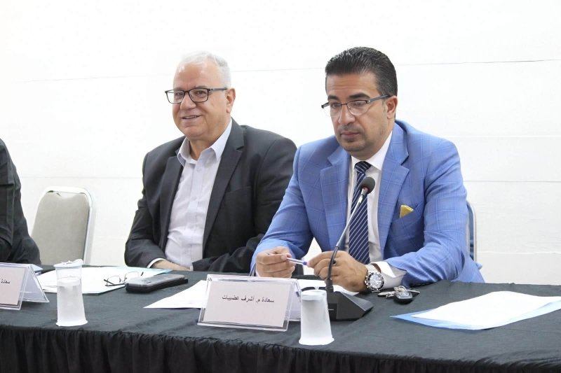 6a74778be7eb9 - عروبة الاخباري | Oroba News Two men in suits sit at a conference table; the man in the blue suit speaks into a microphone beside another man, with name placards and papers in front of them.