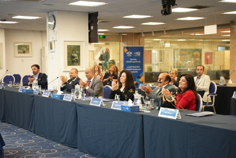 6a772a351c3dc - عروبة الاخباري | Oroba News Panel of professionals seated at a long conference table, clapping during a formal meeting in a bright room with blue tablecloths.