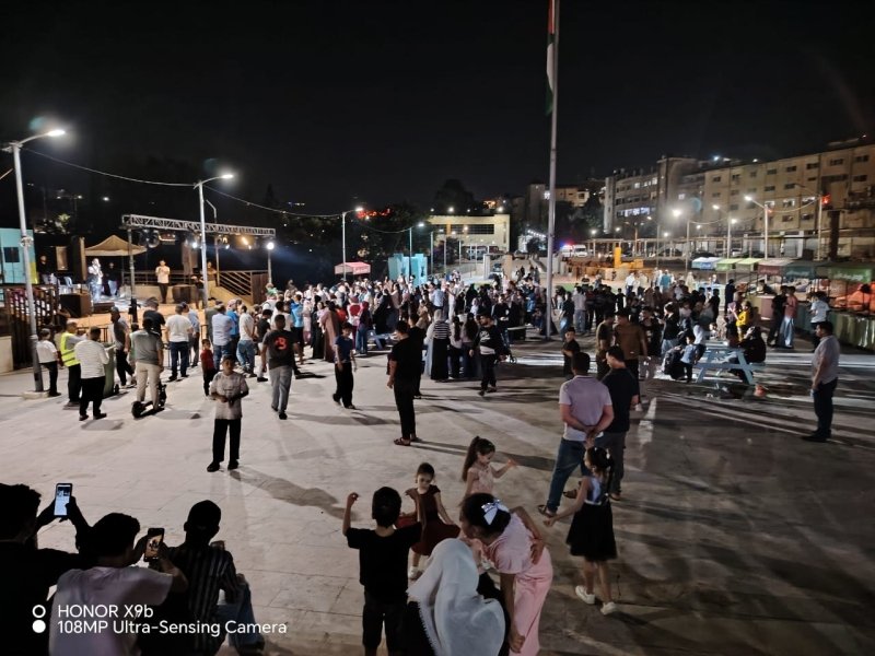 Nighttime outdoor festival in a city plaza with a large crowd, a stage on the left, string lights, and a tall flagpole in the center.