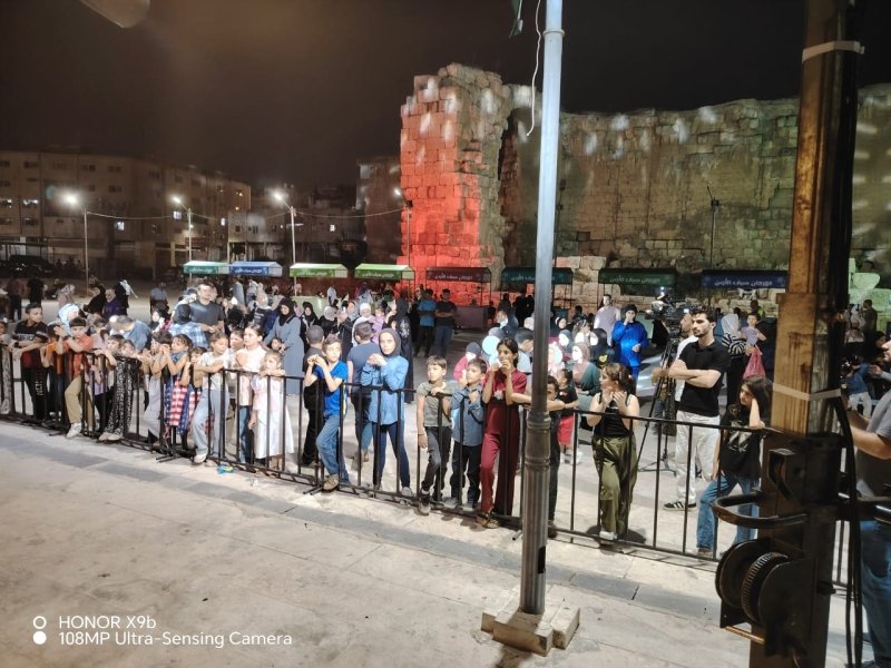 Children standing behind a metal barrier at night near ancient stone ruins illuminated by warm lights.