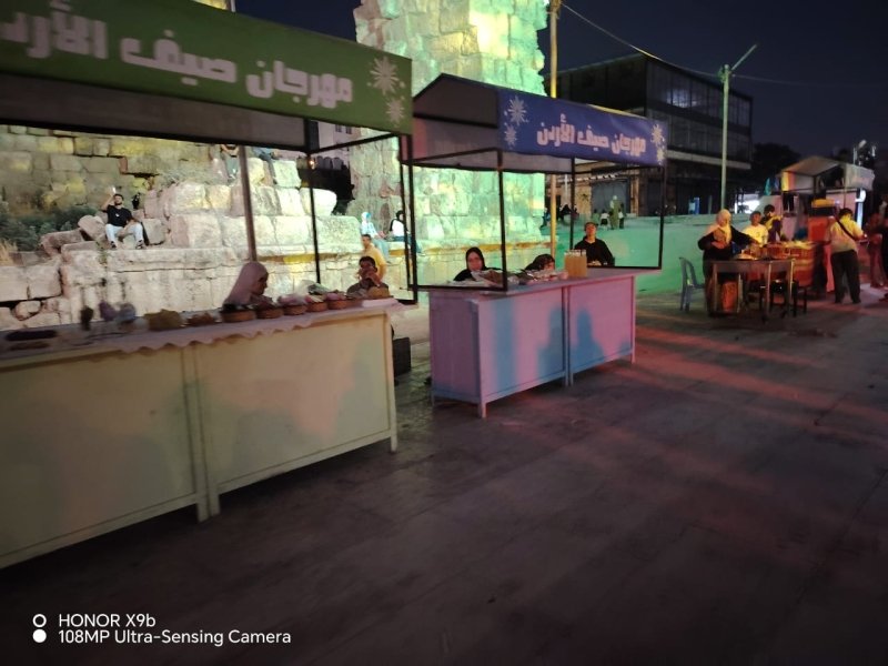Night market stalls with vendors selling goods along a stone wall under green and blue canopies at dusk