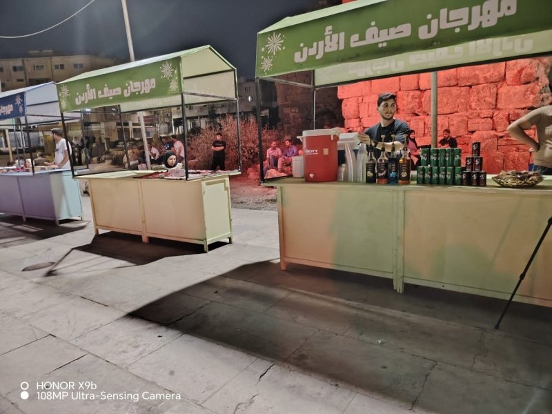 Outdoor night market with green canopies; beverage stand on the right and other vendor stalls in a row behind.