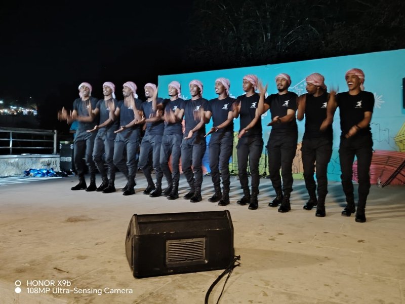 Line of male dancers in matching outfits and head wraps perform in a row on an outdoor night stage, with a speaker placed at the front.