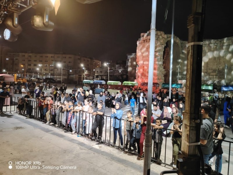 Crowd of people, including children, standing behind a metal barrier at a night outdoor event with lights and vendor stalls in the background.