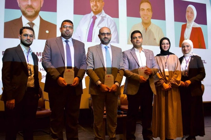 6a905c9c51f57 - عروبة الاخباري | Oroba News Six professionals on stage at an awards ceremony, each holding a plaque, with a backdrop of large portrait panels.