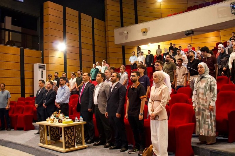 6a905c9c6bfd2 - عروبة الاخباري | Oroba News People standing in a formal auditorium with red seats; a front table decorated with flowers and drinks sits near the stage.