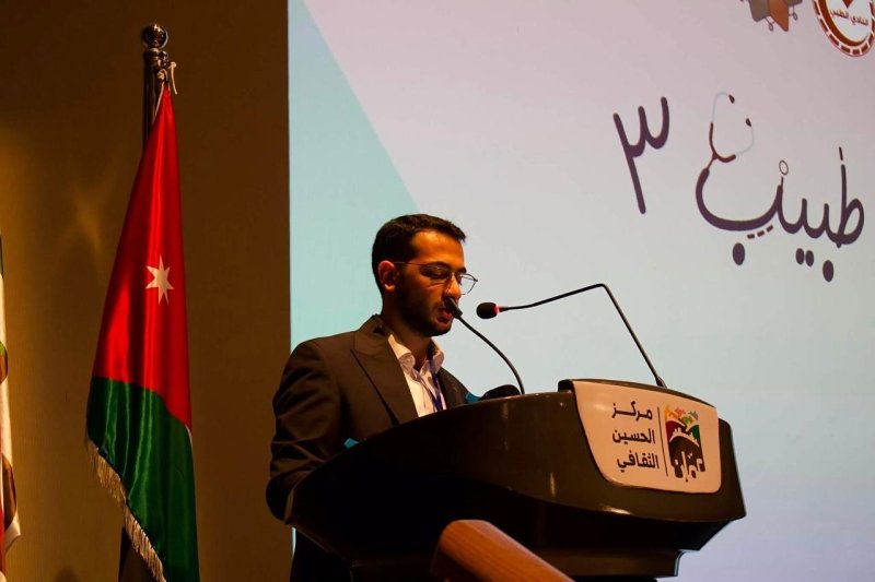 6a905c9c79db9 - عروبة الاخباري | Oroba News Man in a suit delivering a speech at a podium, with a Jordan flag beside him during a conference show.