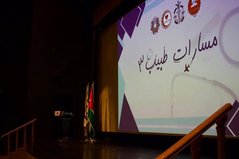 6a905c9c8540c - عروبة الاخباري | Oroba News Stage with a large projection screen displaying Arabic text and geometric designs; two flags stand to the left beside a podium on the darkened stage.