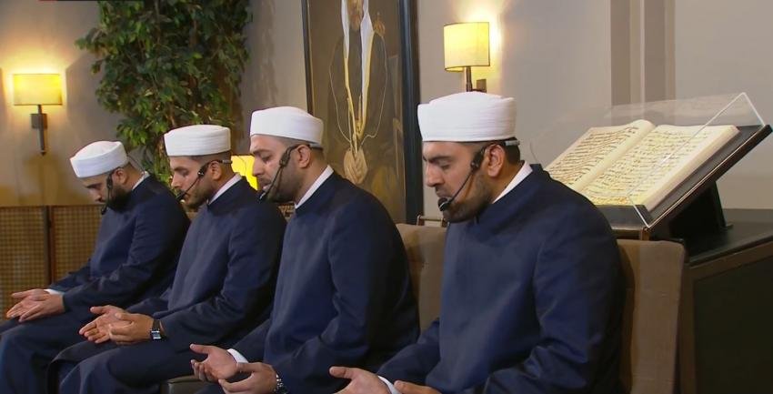 Four men in white turbans and dark robes sit in a row, cupping their hands in prayer in a mosque-ish room with an open book on display behind them.
