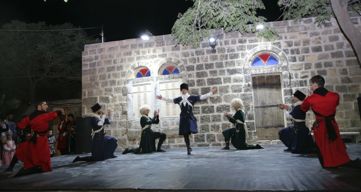 759332 Nighttime outdoor performance on a stone building set, with dancers in traditional costumes (red, blue, white) mid-dance as others kneel and clap nearby, audience figures in background.