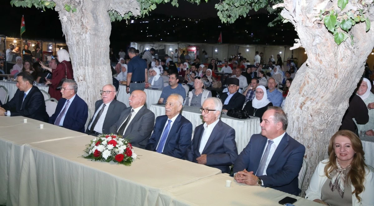 759333 Formal outdoor event with a row of dignitaries seated at a long table topped with a flower centerpiece, under large trees, while a crowd watches from behind.