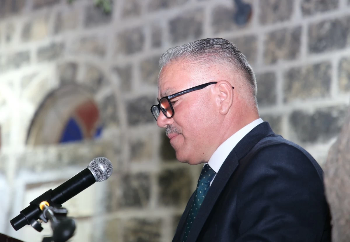 759335 Man in a dark suit and glasses speaks into a microphone at a podium, standing against a stone wall background.