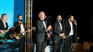 Group of male musicians in suits performing live on an outdoor stage at night, singing into microphones with guitarists and drums in the background.