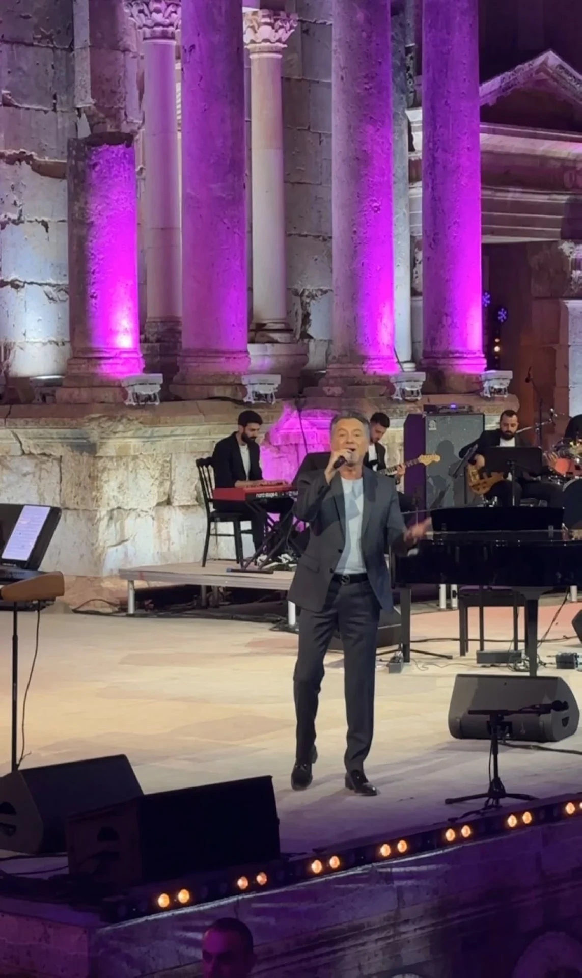 759618 Male singer in a gray suit performing on an outdoor stage with purple-lit ancient columns behind him.