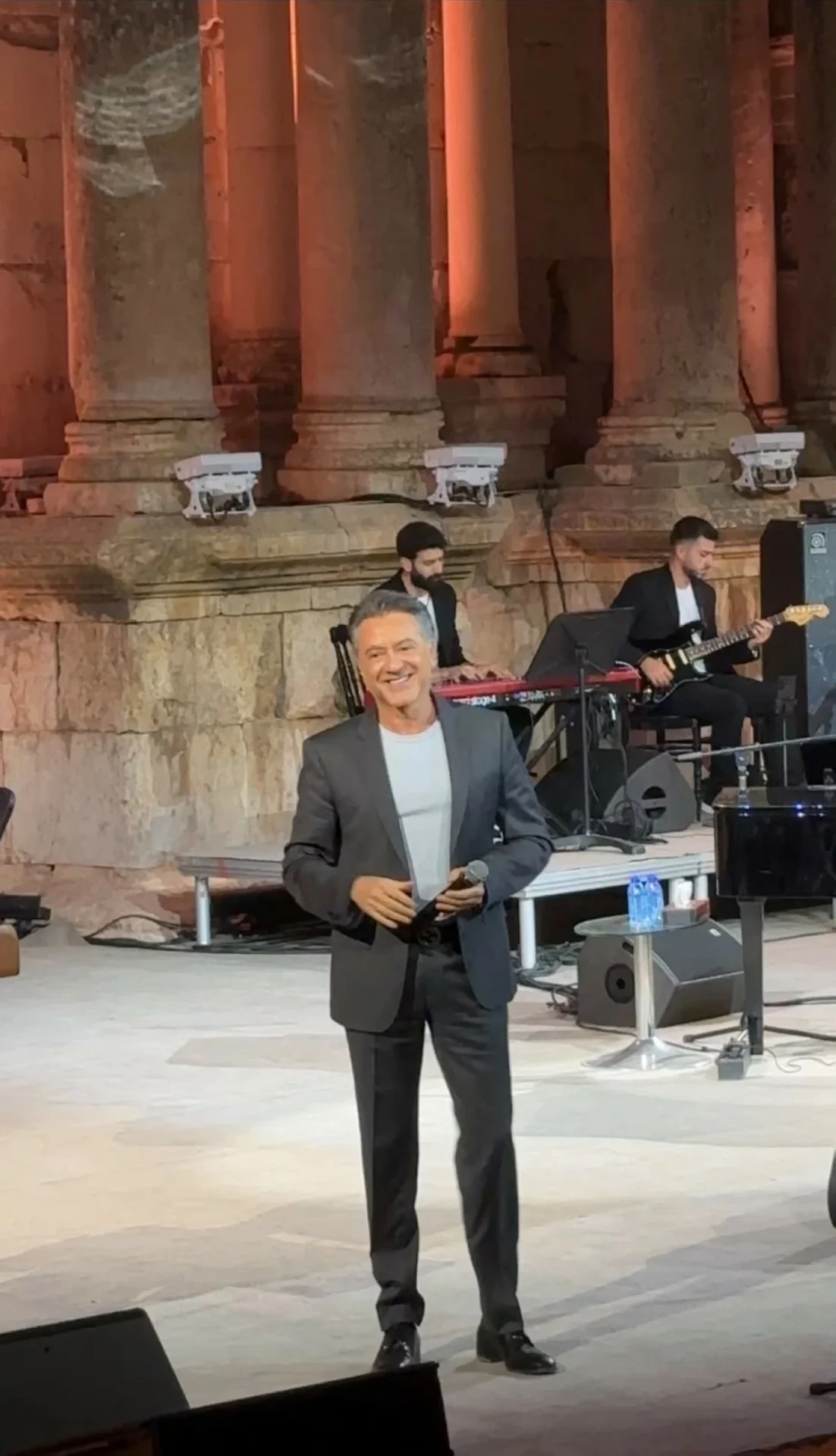 759619 Smiling male singer in a gray suit holding a microphone on an outdoor stage, with a live band (keyboard and guitar) and ancient stone columns in the background.