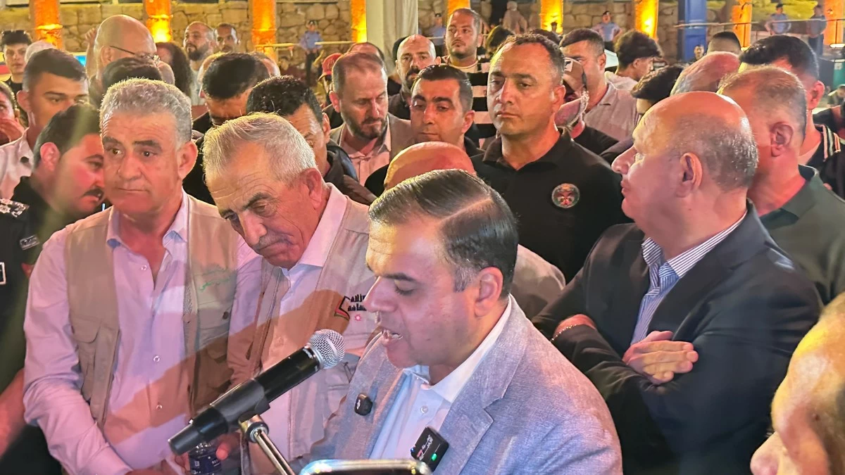 759845 Man in a light gray blazer speaks into a microphone at a crowded indoor event, surrounded by attentive men.