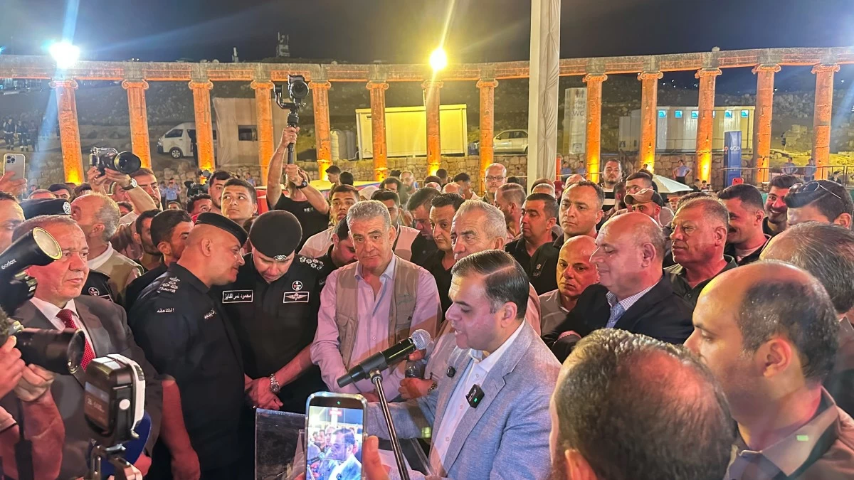 759846 Speaker in a light blazer addresses a crowded audience at night under orange-lit stone columns; reporters with cameras surround the microphone podium.